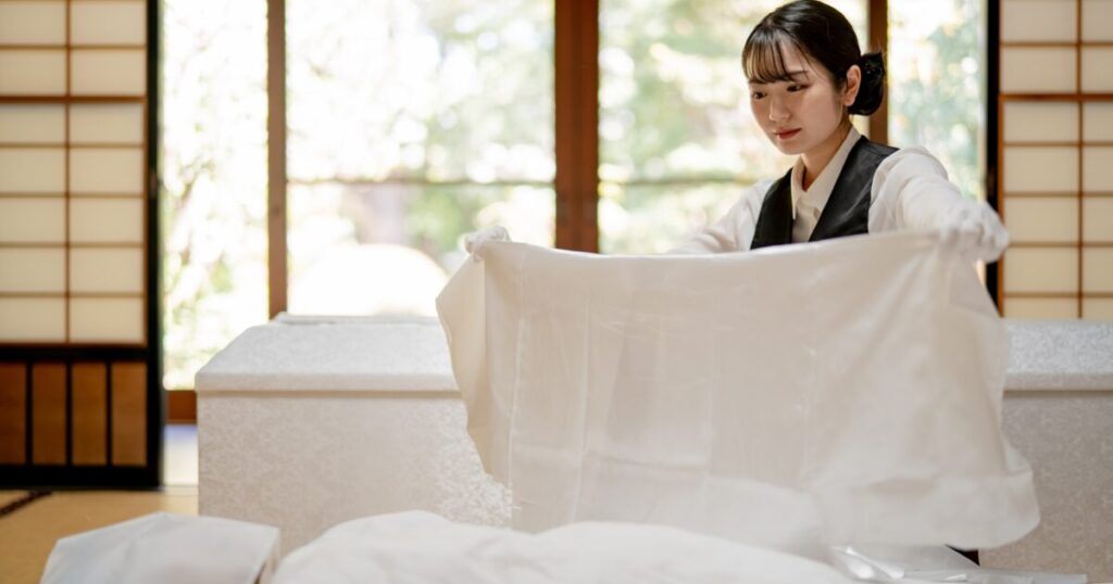 若い女性の葬儀スタッフが丁寧に白い布を広げて納棺の準備をしている様子